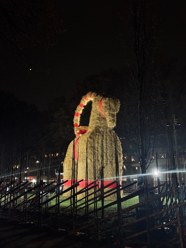 En imponerade syn av Gävlebocken i nattbelysning. Skulpturen är gjorde av halm och smyckad med röda banda, stående bakom ett staket med byggnader och träd i de mörka bakgrunden